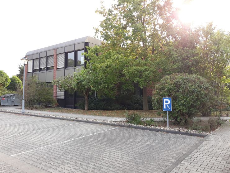 Ein modernes Gebäude mit großen Fenstern, umgeben von Grünflächen, mit einem Parkplatz davor.