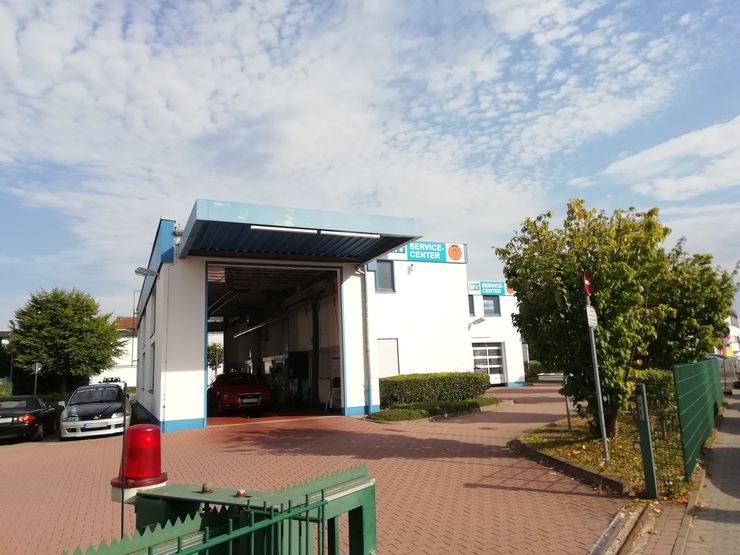 Ein weißes Gebäude mit blauem Dach mit der Aufschrift "TUEV Hessen Service Center", in dem Autos geparkt sind. Im Vordergrund befinden sich eine rote Ampel und ein grüner Zaun.