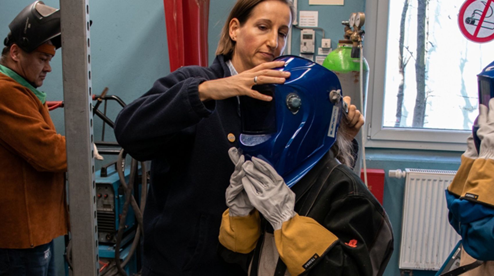 Eine Frau setzt einer Person einen blauen Schweißerhelm auf den Kopf, während eine andere Person in einer braunen Jacke in der Nähe arbeitet.