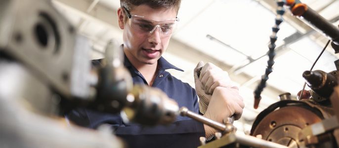 Ein Mann mit Schutzbrille und Handschuhen arbeitet an einer Maschine in einer Werkstatt.