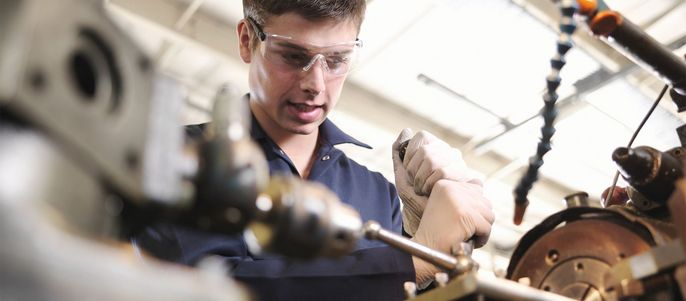 Ein Mann mit Schutzbrille und Handschuhen arbeitet an einer Maschine in einer Werkstatt.
