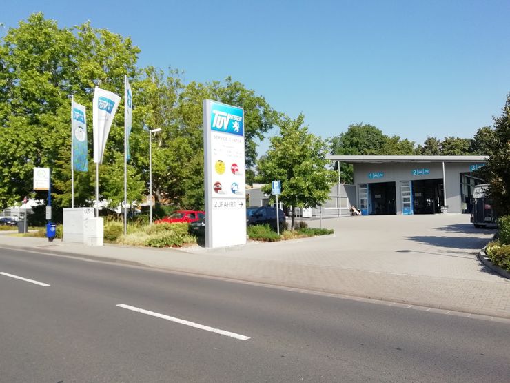 Eine moderne Tankstelle mit einem blau-weißem TUEV Hessen Schild, mehreren geparkten Autos und ein paar Bäumen im Hintergrund.