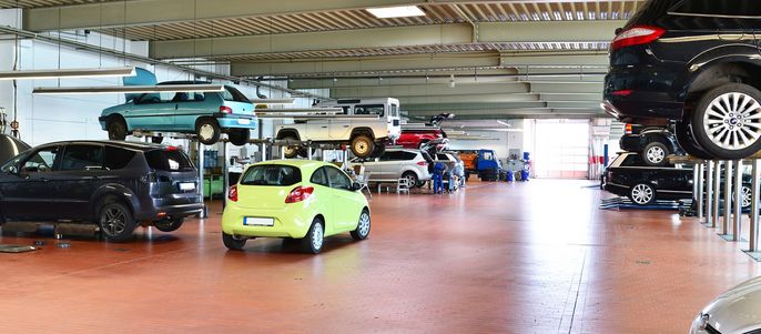 Eine geräumige Garage mit verschiedenen Autos auf Hebebühnen, verstreutem Werkzeug und einigen Arbeitern, die sich um die Fahrzeuge kümmern.