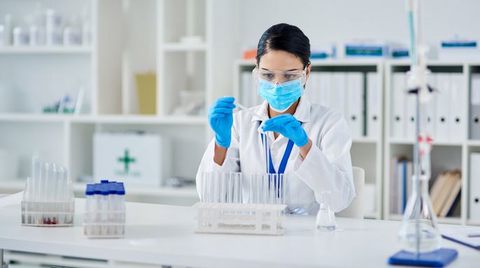 Eine Frau in einem Laborkittel und blauen Handschuhen arbeitet mit einem Reagenzglas.