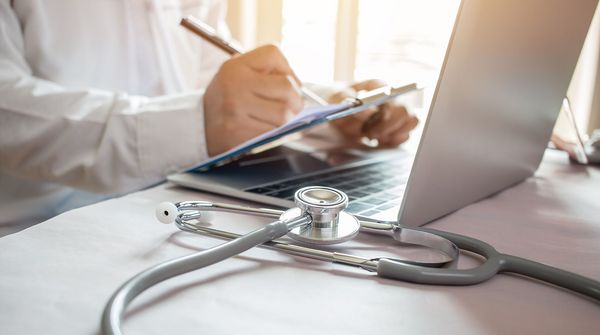 Arzt schreibend vor Laptop und Stethoskop
