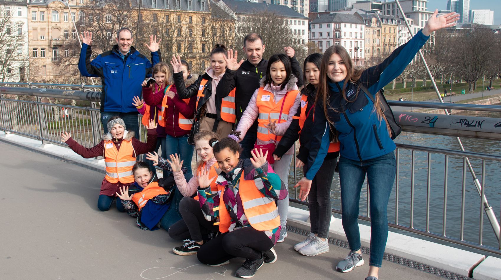 Eine Gruppe von Menschen, sowohl Erwachsene als auch Kinder, stehen und knien auf einer Brücke, tragen leuchtend orangefarbene Westen und winken der Kamera zu.