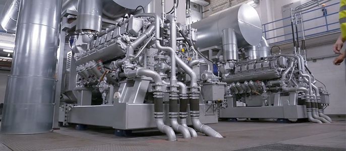 Eine große Industriemaschine, insbesondere ein Motor, mit mehreren Rohren und Bauteilen, in einem Raum mit metallischen Strukturen und einer Person in gelber Weste im Hintergrund.