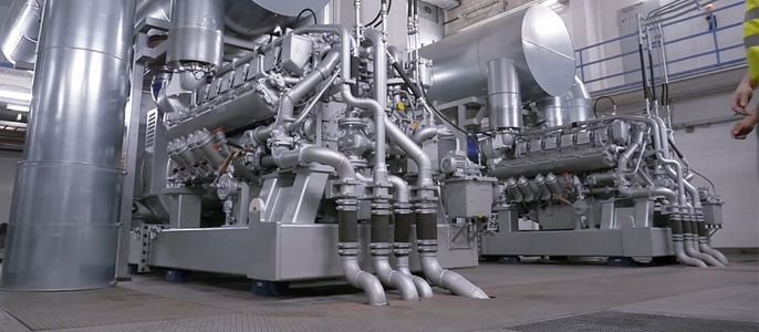 Eine große Industriemaschine, insbesondere ein Motor, mit mehreren Rohren und Bauteilen, in einem Raum mit metallischen Strukturen und einer Person in gelber Weste im Hintergrund.