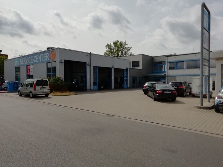 Ein Service-Center mit einem blauen Schild mit der Aufschrift SERVICE-CENTER", ein weißer Lieferwagen, der vor dem Gebäude geparkt ist, und mehrere Autos auf dem Parkplatz.