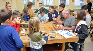Eine Gruppe von Kindern hat sich in einem Klassenzimmer um einen Tisch versammelt und ist mit Werkzeugen und Materialien beschäftigt.