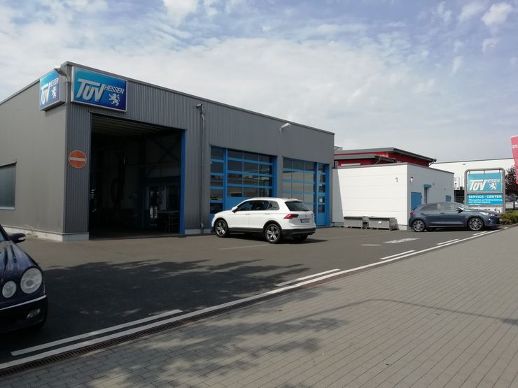 Eine Autowerkstatt mit einem blauen Schild mit der Aufschrift "TUEV Hessen", einem weißen Auto, das drinnen geparkt ist, und ein paar anderen Autos draußen.