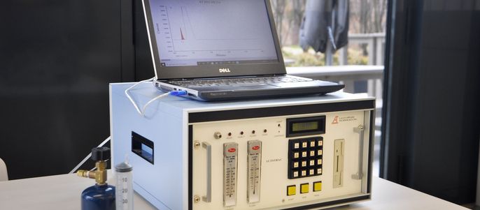 Eine hochentwickelte Maschine mit einem Laptop oben drauf, der mit verschiedenen Schläuchen und Knöpfen verbunden ist. Daneben stehen ein blauer Zylinder und eine durchsichtige Flasche.