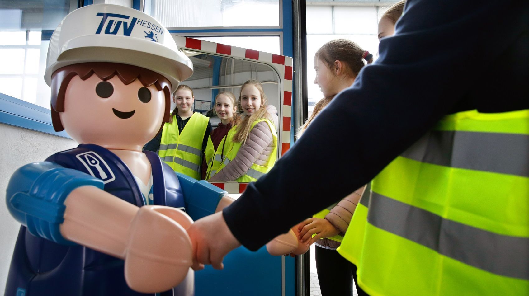 Eine große Playmobil-Figur, die als TUEV Hessen Mitarbeiter gekleidet ist und einer Person, die eine Warnweste trägt, die Hand schüttelt.