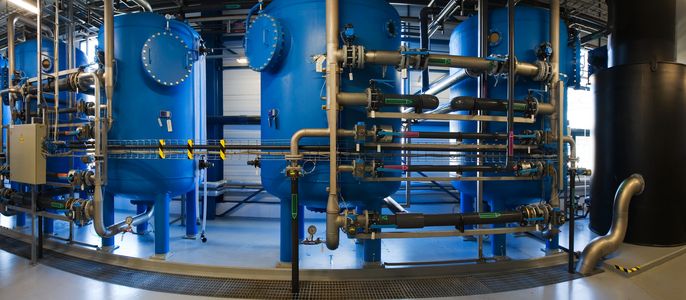 Eine große Industrieanlage mit blauen zylindrischen Tanks, die durch Metallrohre verbunden sind und sich in einem Raum mit reflektierendem Boden befinden.