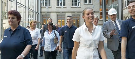 Eine Gruppe von Menschen, die gemeinsam in einem modernen Gebäude spazieren gehen, einige von ihnen tragen Uniformen mit dem Logo "TUEV Hessen".