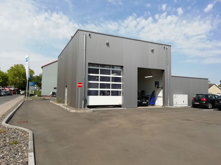 Ein modernes graues Gebäude mit einem Garagentor, das auf einem gepflasterten Grundstück steht und in dessen Nähe ein Auto geparkt ist.