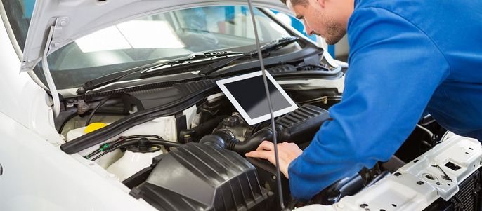 Ein Mann in blauer Uniform arbeitet am Motor eines Autos, auf dessen Motorhaube ein Tablet mit Informationen angebracht ist.