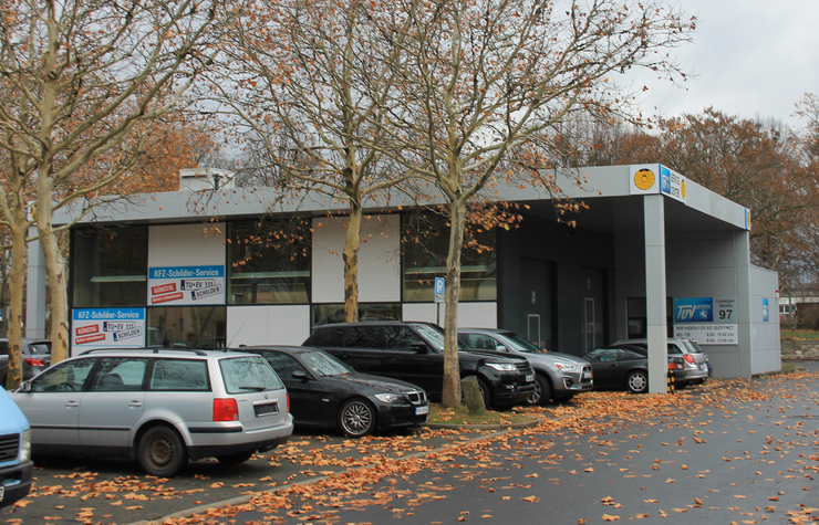 Ein Autohaus mit mehreren geparkten Autos, ein Gebäude mit dem Schild "KFZ-Schulden-Service" und ein Werbebanner für einen Autoverkauf.