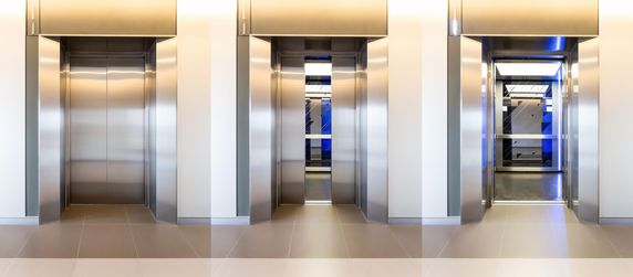 Drei Aufzugstüren, von denen die mittlere geöffnet ist und einen blau beleuchteten Innenraum freigibt.