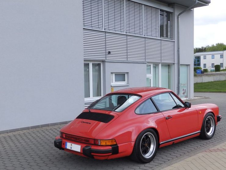 Ein roter Porsche parkt vor einem Gebäude mit weißer Fassade und schwarzem Fenster.
