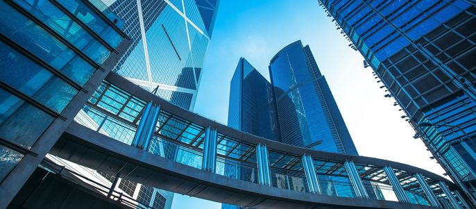 Moderne Wolkenkratzer mit spiegelnden Glasfassaden, die durch einen transparenten Gehweg miteinander verbunden sind, vor einem strahlend blauen Himmel.