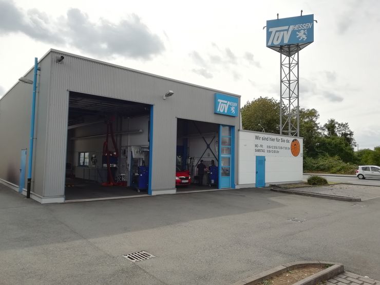 Eine Autowerkstatt mit dem Namen "TUEV Hessen", mit einem Schild auf einem Turm, einem Auto in der Werkstatt und ein paar anderen Fahrzeugen draußen.
