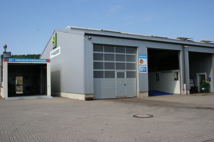 Ein Gebäude mit einem Schild mit der Aufschrift "WAGNER" und einem weiteren Schild mit der Aufschrift "ABGASUNTERSUCHUNG". Es gibt zwei Garagentore, von denen eines geöffnet ist und den Blick auf eine Autowaschanlage freigibt.