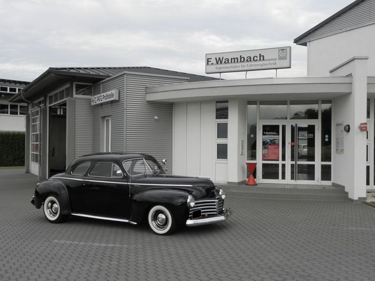 Ein schwarzer Oldtimer parkt vor einem modernen Gebäude mit der Aufschrift "F. Wambach".