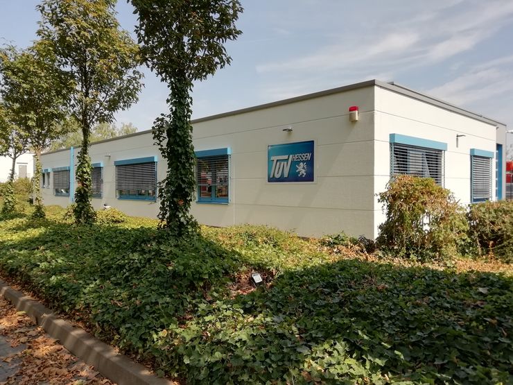 Ein weißes Gebäude mit einem blauen Schild mit der Aufschrift "TUEV Hessen". Das Gebäude hat Fenster mit Jalousien, und es ist von Bäumen und Grün umgeben.
