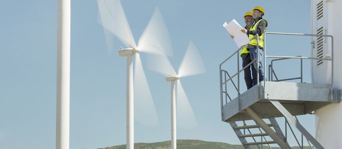 Zwei Arbeiter auf einer Plattform in der Nähe von Windkraftanlagen, einer von ihnen hält einen Bauplan.