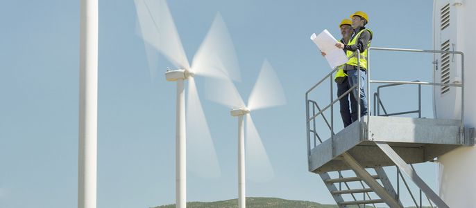 Zwei Arbeiter auf einer Plattform in der Nähe von Windkraftanlagen, einer von ihnen hält einen Bauplan.