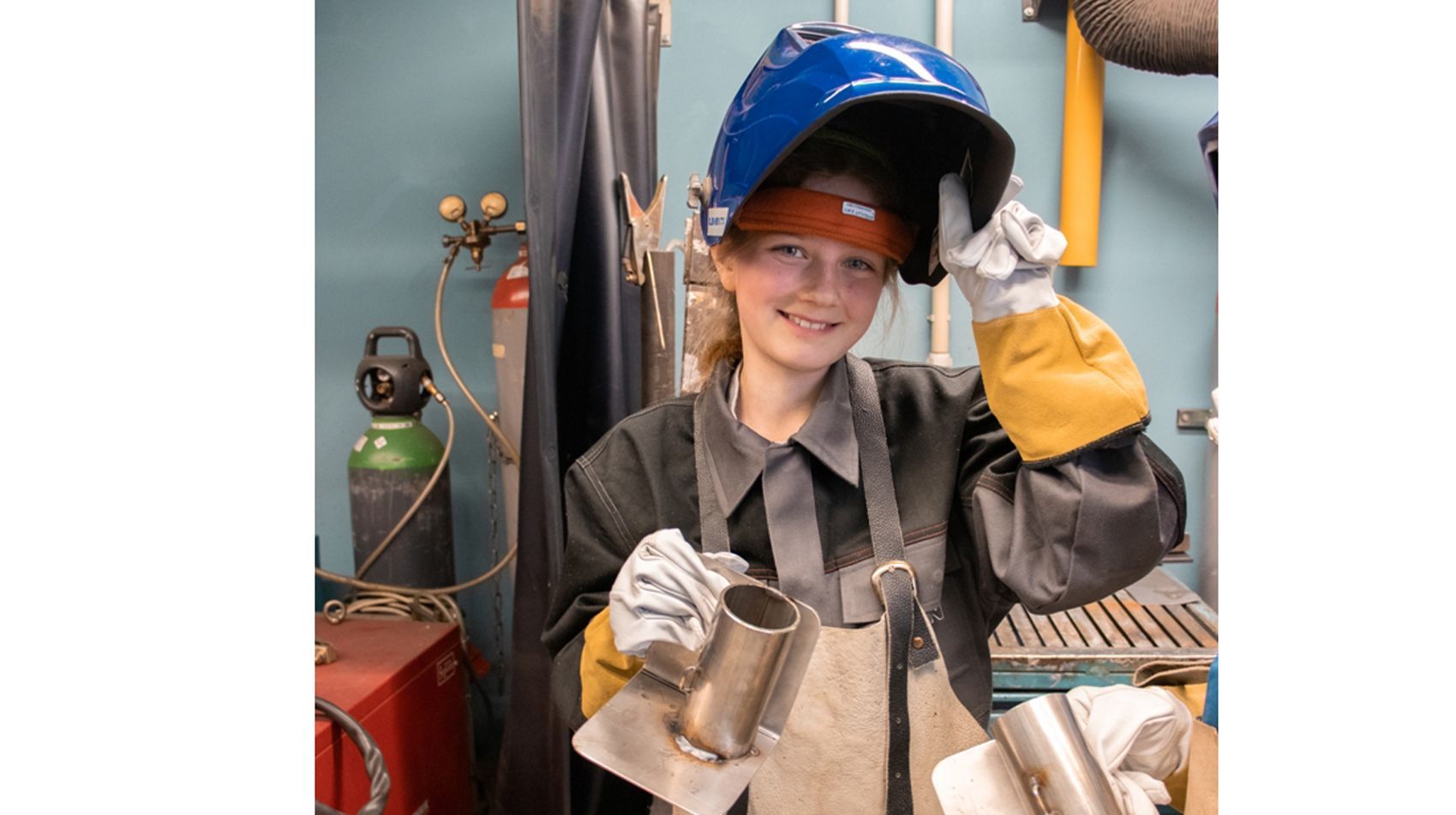 Eine junge Frau in einer Werkstatt, mit Schutzkleidung, die einen Metallzylinder und ein Schweißgerät hält.