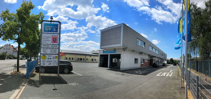 Ein modernes Gebäude mit einem Schild mit der Aufschrift "TUEV Hessen" und "Technische Dienstleistungen", umgeben von einem Parkplatz mit Autos und einem blauen Zaun.
