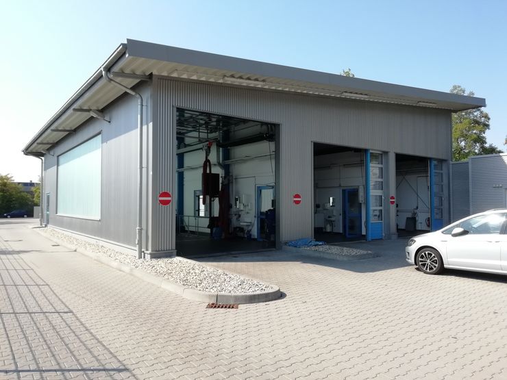 Ein modernes Industriegebäude mit grauem Äußeren, ausgestattet mit großen Türen, einem weißen Auto, das draußen parkt, und verschiedenen Maschinen im Inneren.