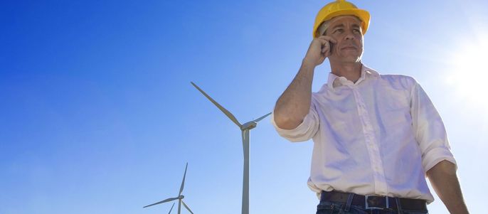 Ein Mann in einem weißen Hemd und einem gelben Schutzhelm spricht mit einem Mobiltelefon vor dem Hintergrund von Windkraftanlagen unter einem klaren blauen Himmel.