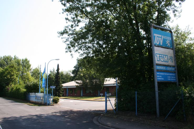 Ein Schild für "TUEV Hessen" mit dem Untertext "SERVICE-CENTER", umgeben von Bäumen und einem Gebäude.