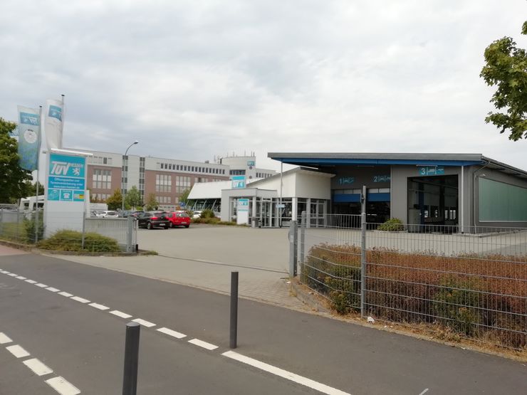 Ein modernes Gebäude mit blau-weißer Fassade, einem Schild mit der Aufschrift "TUEV Hessen" und ein paar geparkten Autos.