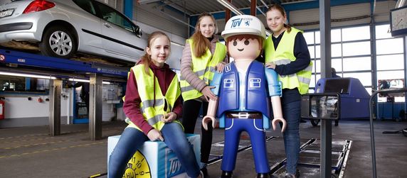 Vier Personen in einer Werkstatt, in der Mitte eine große Playmobil-Figur, die als TUEV Mechaniker verkleidet ist.