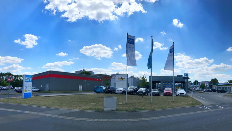 Ein modernes Autohaus mit mehreren draußen geparkten Autos, einigen Fahnen und einem Gebäude mit dem TUEV Hessen-Logo.