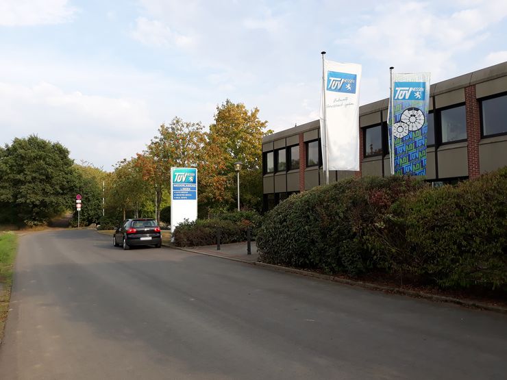 Eine Straße, die zu einem Gebäude mit dem "TUEV Hessen"-Logo führt, umgeben von Bäumen und Sträuchern, an dessen Seite ein Auto geparkt ist.