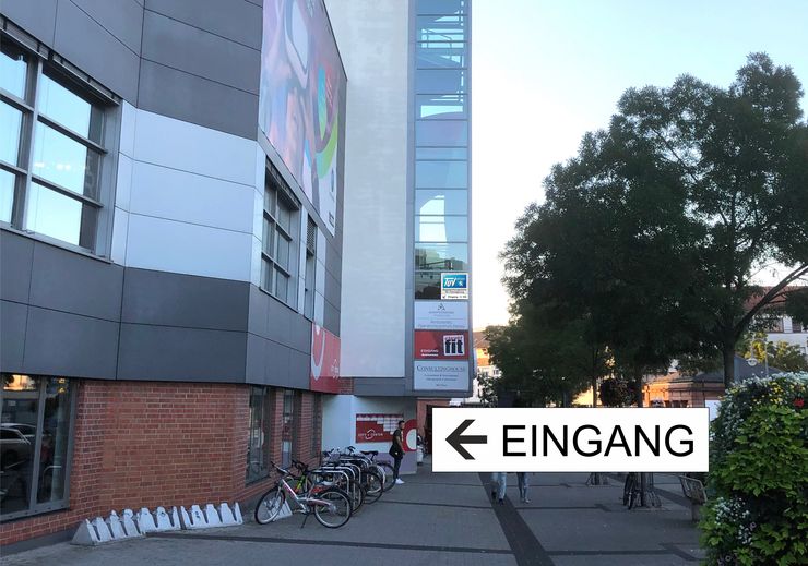 Ein modernes Gebäude mit einer großen Glasfassade, einem Schild, das nach links auf eine Hausöffnung weist, und ein paar Fahrrädern, die draußen abgestellt sind.