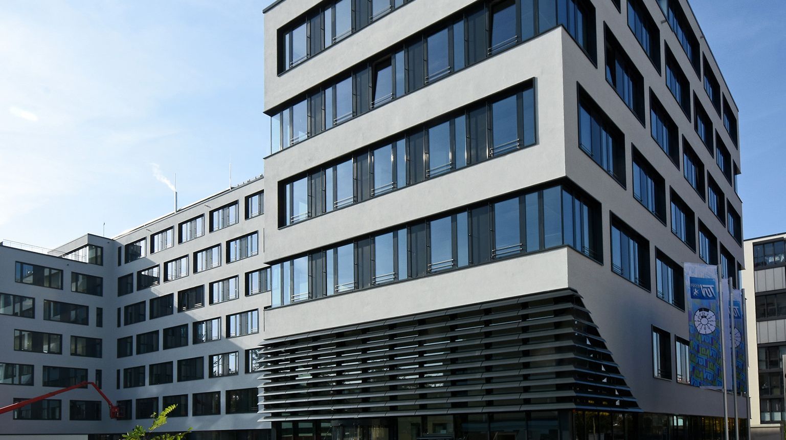 Ein modernes mehrstöckiges Gebäude mit überwiegend weißer Fassade, zahlreichen Fenstern und einem markanten schwarzen Gitter im unteren Bereich. Das Gebäude befindet sich in einer städtischen Umgebung mit einem klaren, blauen Himmel über sich.