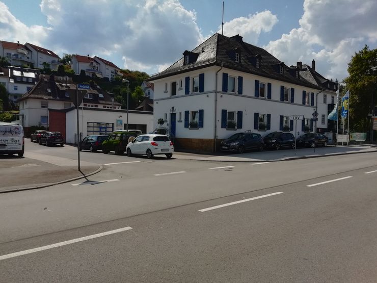 Eine Straßenszene mit einem weißen Gebäude, blauen Fensterläden und geparkten Autos. Links steht ein Wegweiser und rechts ein Gebäude mit einer blauen Flagge.