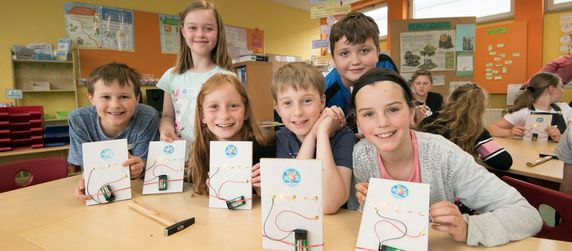 Eine Gruppe von Kindern in einem Klassenzimmer hält Schaltkreise mit dem TUEV Kids Logo und einem kleinen elektronischen Gerät in die Höhe.