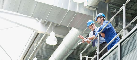 Zwei Männer in blauen Uniformen stehen auf einer Plattform und betrachten ein Dokument in einer großen Industrieanlage mit verschiedenen Rohren und Maschinen.