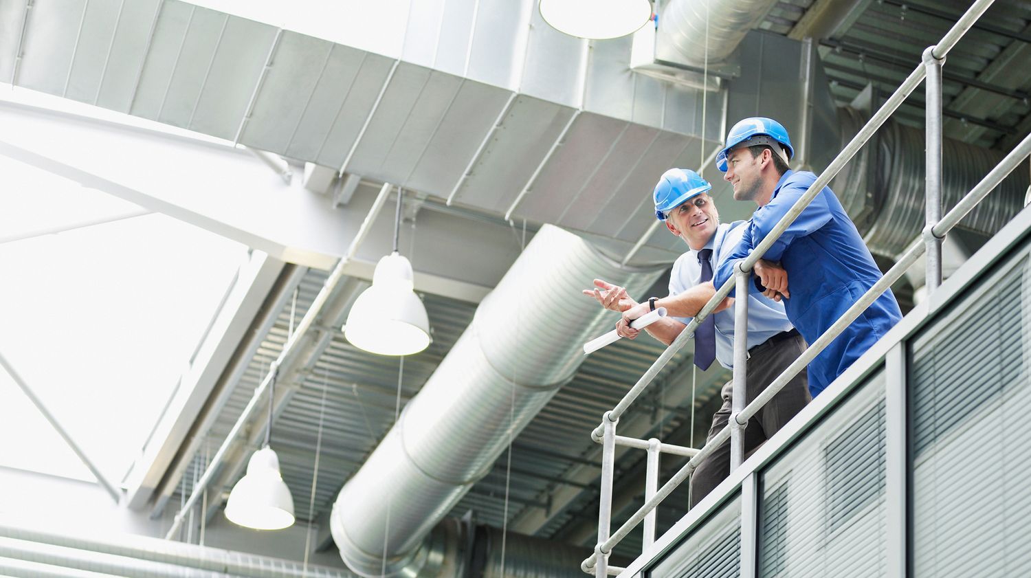Zwei Männer in blauen Uniformen stehen auf einer Plattform und betrachten ein Dokument in einer großen Industrieanlage mit verschiedenen Rohren und Maschinen.