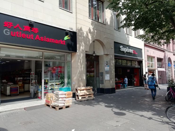 Eine Straßenszene mit zwei Geschäften: "Gutleut Asiamarkt" und "Sapores", beide mit ihrer jeweiligen Beschilderung. Vor dem "Gutleut Asiamarkt" sind Holzpaletten gestapelt, und in der Nähe des "Sapores" ist eine Person zu sehen.