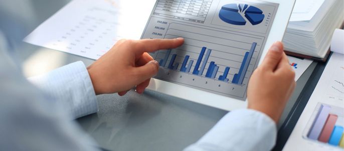 Eine Person, die ein Balkendiagramm auf einem Tablet analysiert, mit verschiedenen Finanzdatenblättern und Diagrammen im Hintergrund.