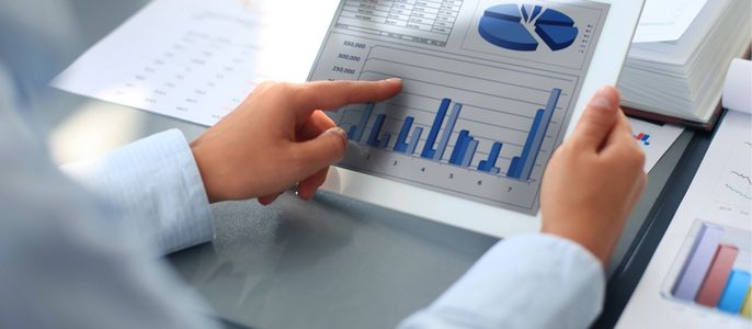 Eine Person, die ein Balkendiagramm auf einem Tablet analysiert, mit verschiedenen Finanzdatenblättern und Diagrammen im Hintergrund.