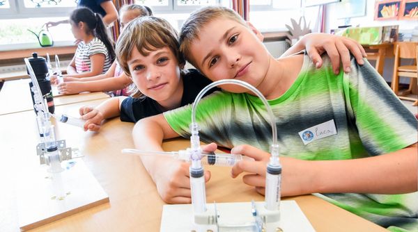 Zwei Jungen in einem Klassenzimmer, beide lächeln und schauen in die Kamera. Sie halten eine TUEV Kids Hebebühne in der Hand.